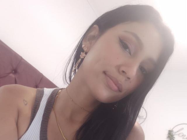KataleyaMansson has long black hair, brown eyes, and a small nose tattoo. She's wearing a white tank top, gold necklace, and ornate earrings, smiling with her head tilted.