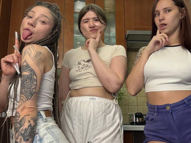 Three HotCaramelsTrio standing in a kitchen, wearing white tops and casual pants. Left has braids and tattoos, middle has short brown hair, right has long brown hair. Sticking tongues out.