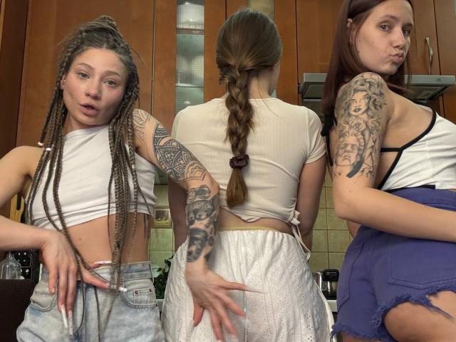Three HotCaramelsTrio standing in a kitchen. One with long braids and tattoos, another with a braid, and the third with tattoos and a purple skirt, all showing their asses.