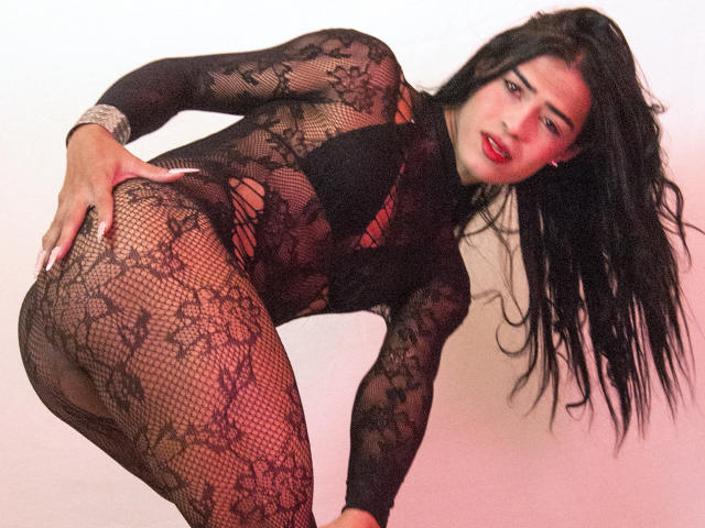 VioletSexyHot met lang zwart haar, rode lippenstift, gekleed in een zwart kanten body, gebogen, hand op haar billen, waarbij haar achterwerk zichtbaar is door het gaas, kijkend naar de camera..