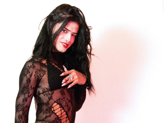 VioletSexyHot mit langen schwarzen Haaren, trägt ein durchsichtiges schwarzes Spitzenkleid und zeigt ihre Brüste, wobei sie einen Ring am Hand trägt und leicht lächelt..
