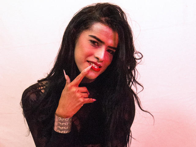 VioletSexyHot, con lunghi capelli neri, rossetto rosso e un sorriso malizioso, indica le sue labbra rosse con un dito, indossando una maglia nera in rete e un braccialetto d'argento..