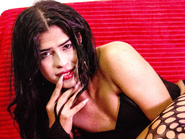 VioletSexyHot, con capelli scuri, labbra rosse, un abito nero e maniche in rete, che tocca le labbra con un dito, sdraiata su un divano rosso. Il suo seno è parzialmente visibile..