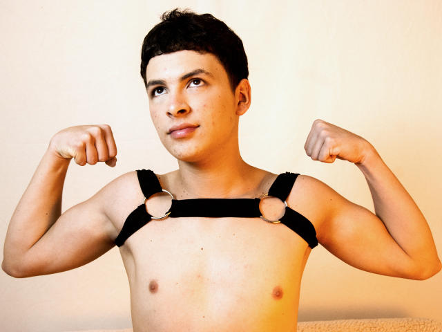 AxelLaciel, a cute young guy with short dark hair, flexes his muscular arms while wearing a black leather harness. His nipples are visible. He has a slight smile.
