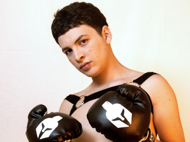 AxelLaciel with short curly black hair, wearing black boxing gloves with white emblem, black harness, and looking serious. Boobs are covered by gloves, showing only straps.