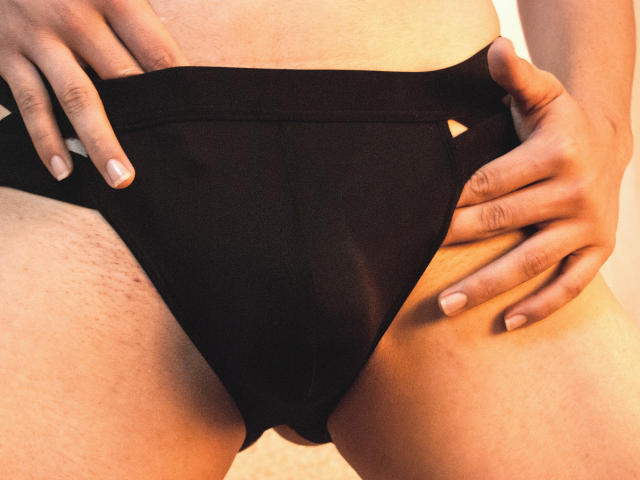 AxelLaciel's hands pull up black panties, exposing a trimmed pussy with visible skin texture. Their fingers have short, clean nails. The focus is on their lower belly and crotch.