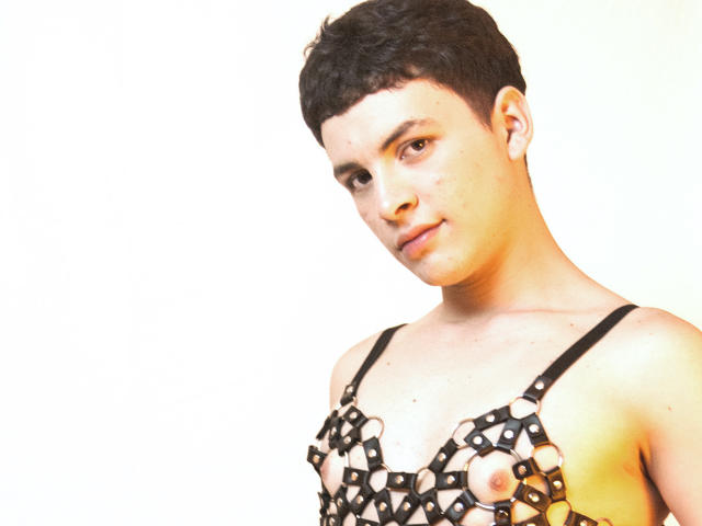 AxelLaciel with short black hair, wearing a black, patterned, cut-out top exposing small, round, brown nipples. Light, slightly yellowed background. Soft, confident expression.