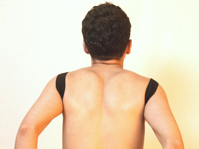AxelLaciel's back is to the camera, shirtless with black straps, showing a muscular, tan butt. He has curly dark hair and is standing against a plain wall.
