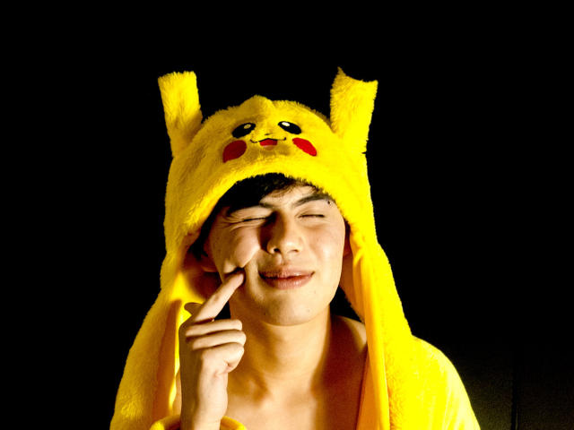 FelipeKold wearing a Pikachu hat, smiling with eyes closed, pointing to his nose, yellow fur hat with Pikachu face, black background, cute, playful expression.