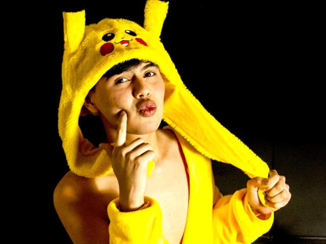 FelipeKold, Asian dude, wearing Pikachu yellow hat and robe, pouting with finger to cheek, yellow fur hat with Pikachu face, black background, illuminated from front.