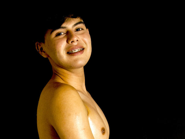 Shirtless FelipeKold with a slight smile, brown short hair, and light skin, showing small nipples and a clean chest against a black background. He's looking over his shoulder.