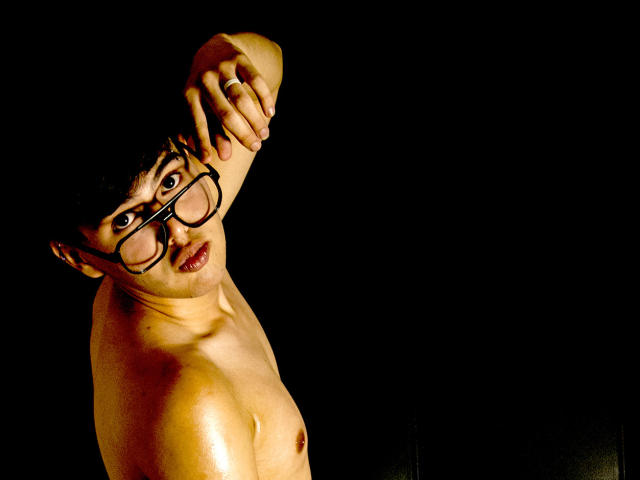 Shirtless FelipeKold with glasses, raising right arm, looking sideways, dark background, visible nipple, slightly chubby, yellow light highlighting upper body, serious expression.