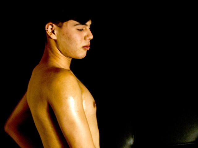  FelipeKold is a shirtless dude with a shiny, sweaty upper body, short dark hair, and a small ear piercing, standing sideways in a dark, black background.