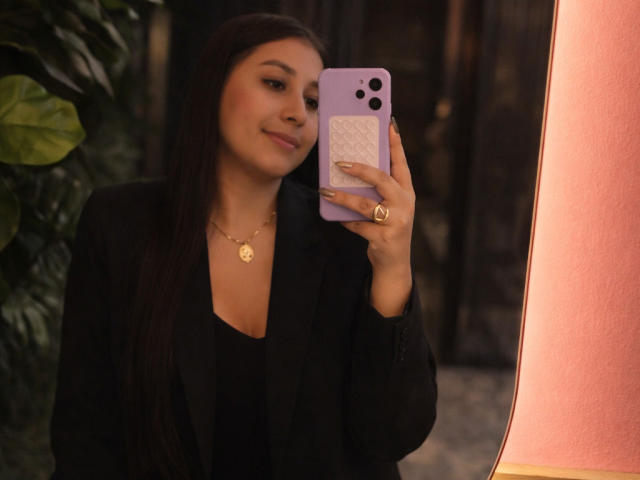 CatalinaRivero with long dark hair, black blazer, and gold necklace, taking a mirror selfie with a purple phone, smiling slightly, showing off her cute face and gold ring.