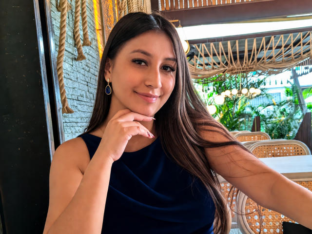 CatalinaRivero with long brown hair, dark blue sleeveless top, smiling, hand on chin, earrings, cute, sexy, tan skin, natural light, wicker chairs, rope decor in the background.
