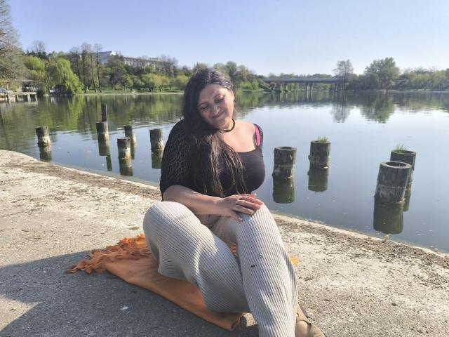 BlissSophia with dark hair, black top, pink bra, and white pants, sitting on an orange blanket by a lake, smiling, with her knees up and hands on her knees.
