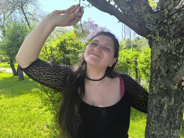 BlissSophia with long black hair, leopard print sleeves, black choker, and pink bra, smiling while holding a leaf from a tree. Her tits are partially visible.