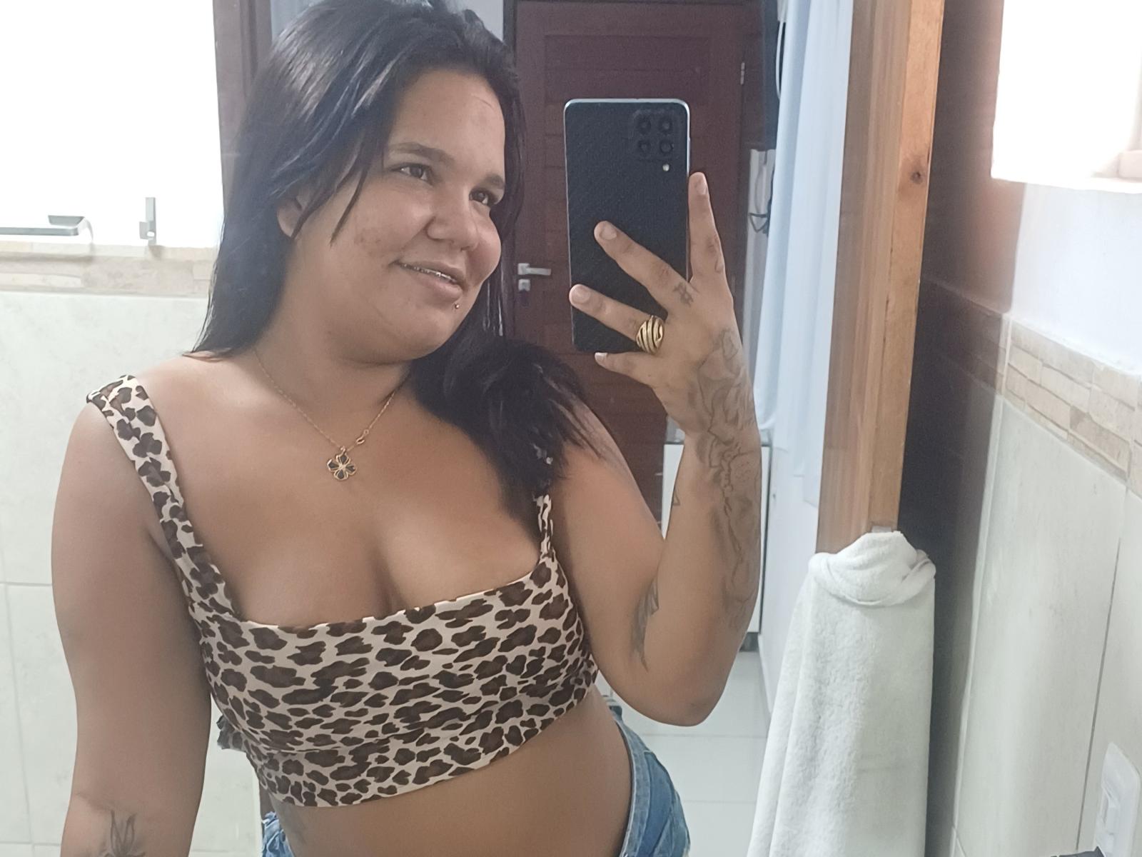 TaniyaS, avec une poitrine de taille moyenne, porte un crop top à motifs léopard, elle sourit, tient un téléphone dans un selfie pris devant un miroir de salle de bain, elle a des tatouages et porte un jean bleu..