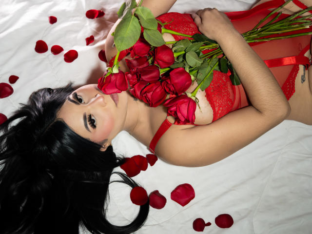 EllieeSmith with black hair, red lace lingerie, holding red roses, petals scattered on white bed, looking up with soft makeup, tattoo on right side of ribcage.