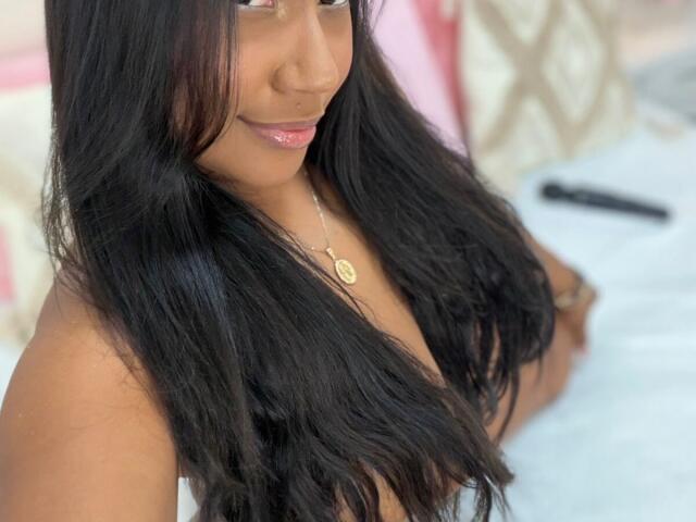 NaiomiLanne with long black hair, dark skin, pink lipstick, gold necklace, and visible large boobs, smiling slightly, on a bed with a white and pink patterned pillow.