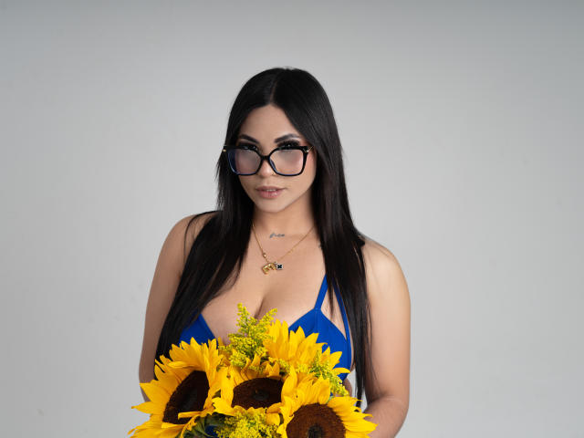 EllieeSmith with long black hair, wearing glasses, blue bikini top, holding sunflowers and yellow flowers, has large tits, light skin, subtle tattoo on neck, gray background.