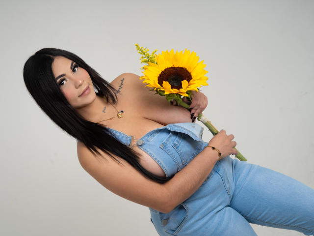 EllieeSmith with long black hair, wearing blue denim overalls, holding a sunflower, showing her big tits, tattoos on her left shoulder, smiling, light skin, in a studio shot.