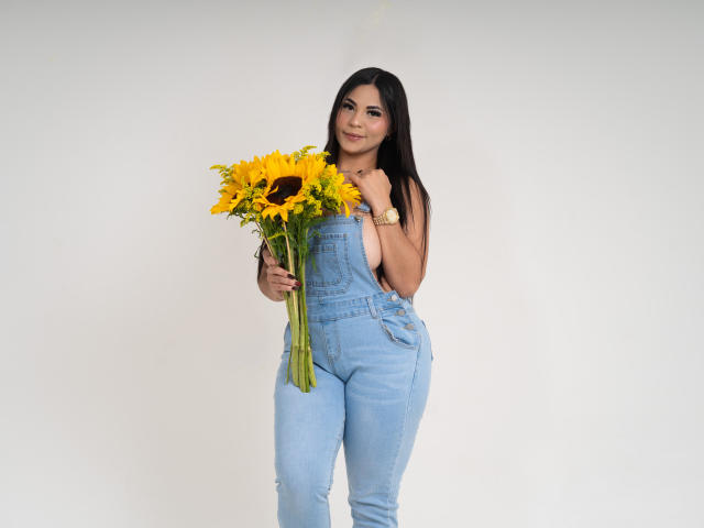 EllieeSmith with long black hair, wearing light blue denim overalls, holding a bouquet of bright yellow sunflowers, standing against a white background, smiling slightly.