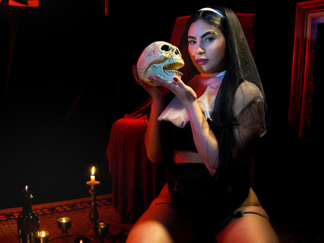 EllieeSmith with long black hair, wearing a sheer black top and veil, holds a skull in her hands, sitting in a dimly lit room with candles and red cloth.