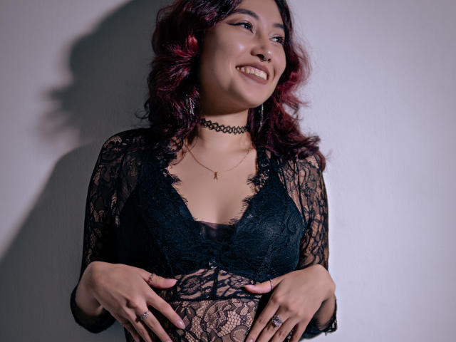 AliCipher with red curly hair, smiling, wearing black lace top, choker, and rings, hands on her tits, gold necklace, soft light, shadow on wall.