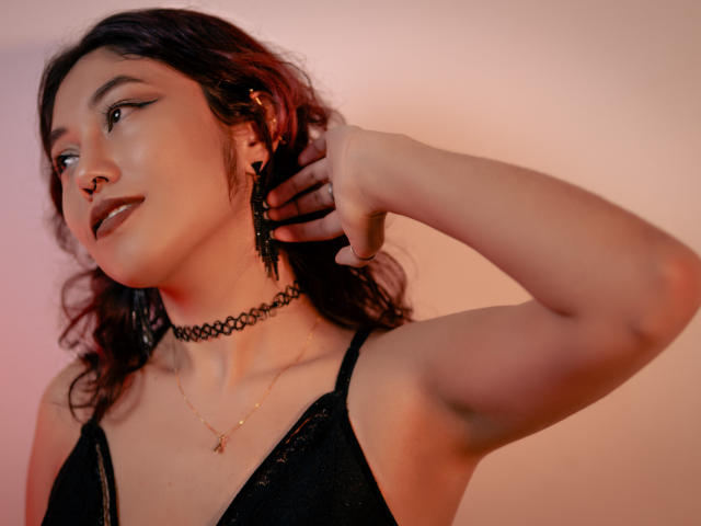 AliCipher has dark wavy hair, black eyeliner, a nose ring, and a black choker. She's adjusting a black earring, wearing a black lace top, with a gold necklace.