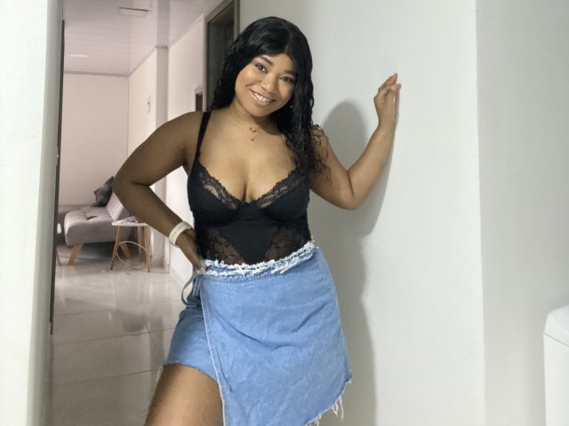 AvaJones69 with dark wavy hair, black lace top, showing her boobs, and blue denim skirt, smiling, hand on white wall, wearing white bracelet, standing in white room.