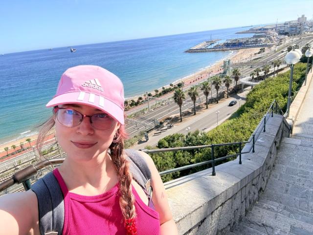 AmbeerWood with pink Adidas cap, pink sports top, glasses, and braid, holding backpack, standing on stone balcony, ocean and beach in background, smiling, showing tits.