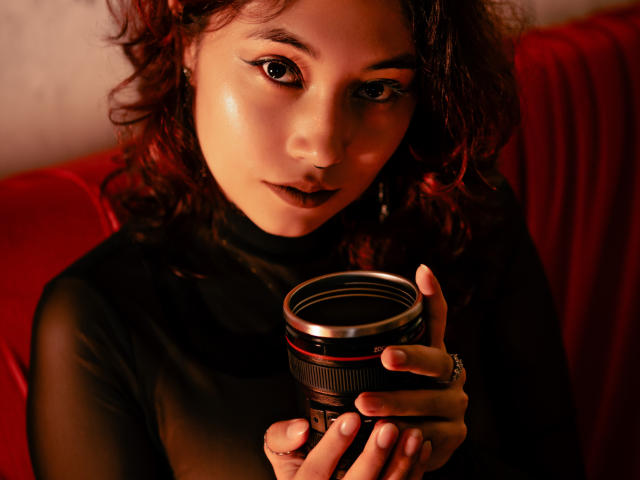 AliCipher, con i capelli rossi ricci, che tiene una macchina fotografica, guarda direttamente l'obiettivo con la pelle luminosa, indossa una maglia nera trasparente, sfondo rosso, espressione seria, primo piano..