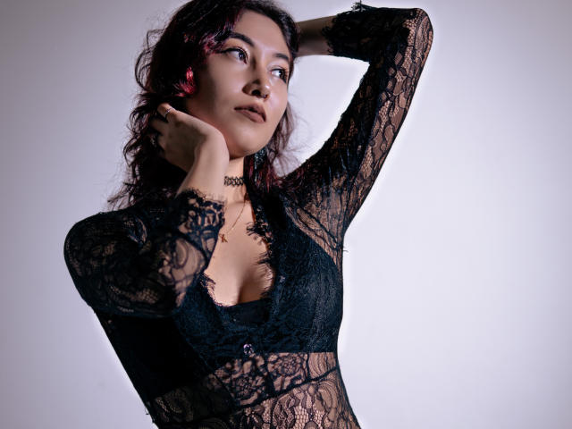 AliCipher with wavy dark hair, wearing a black lace top, has her hands behind her neck, looking to the side, showing her small tits and a black choker.