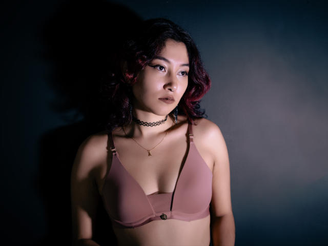 AliCipher with dark curly hair, pink bra, choker, and star necklace, looking serious, light shadows on her, dark background, soft lighting, medium tits, slight tan skin, Asian.