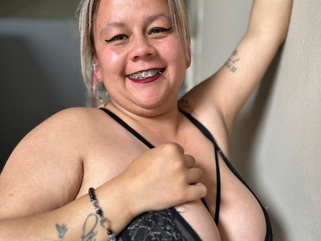ScarletBigTitties, smiling with teeth showing, has gray hair, tattoos on arm and wrist, black lace bra, holding her large tits, standing against a wall.