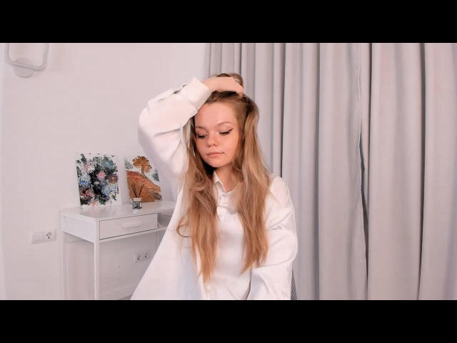 DeliaRodrick with long blonde hair, wearing a white shirt, hand in her hair, looking down, in a softly lit room, with gray curtains and a white table.