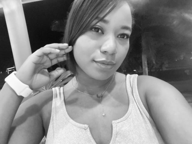 AvaJones69 has light skin, small tits, and a cute face with a slight smile. She's wearing a tank top and a cross necklace, touching her ear. Black-and-white selfie.