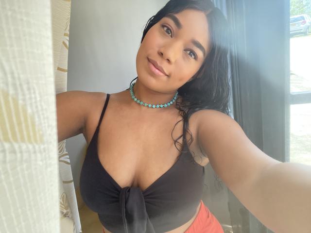 AvaJones69 with dark hair, tan skin, and small nose, wearing a black tied-up top showing her big boobs, and a turquoise bead necklace, smiling at the camera.