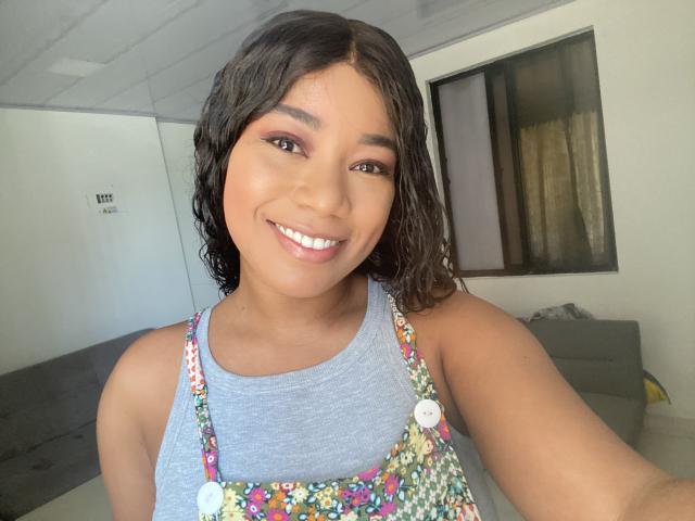 AvaJones69 with curly black hair, brown eyes, and a smile, wearing a floral apron over a grey tank top, showing her cute tits and pretty ass.