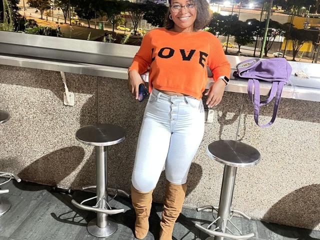 AvaJones69 with curly brown hair, glasses, orange "LOVE" sweater, white jeans, and tan boots, leaning on barstools, smiling, purple bag on bar, daytime, city lights.