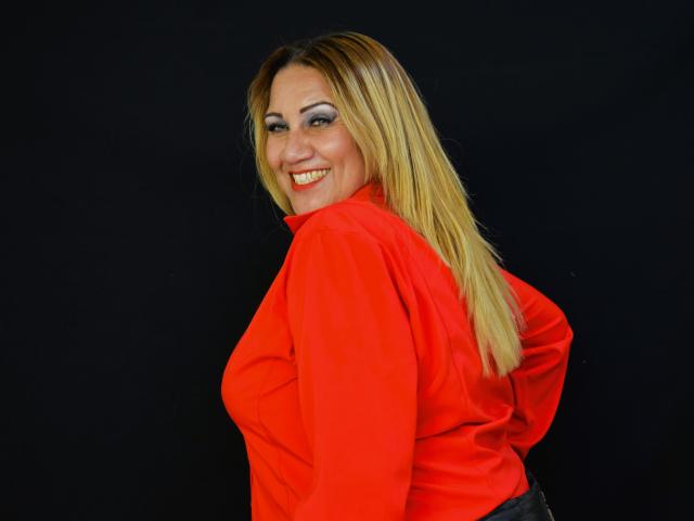 MatureYayita with blonde hair, red shirt, smiling, looking back, showing her tits, black background, wearing makeup, smiling with red lipstick, confident pose.