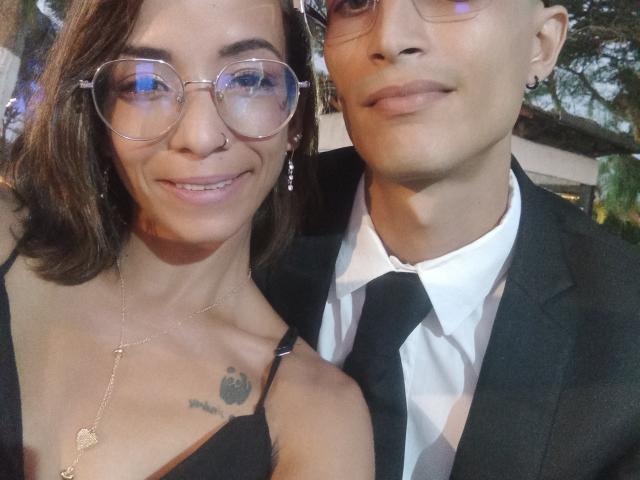 AlejoyMaria with medium tits, black dress, glasses, and necklace, smiling next to a guy in a black suit, white shirt, and black tie. He's wearing glasses too.