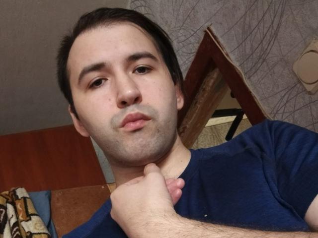 GregArdor mit kurzen, dunklen Haaren, leichtem Bartansatz und einem nachdenklichen Gesichtsausdruck, der seinen Hals mit der rechten Hand hält, ein blaues Hemd trägt und in die Kamera schaut..