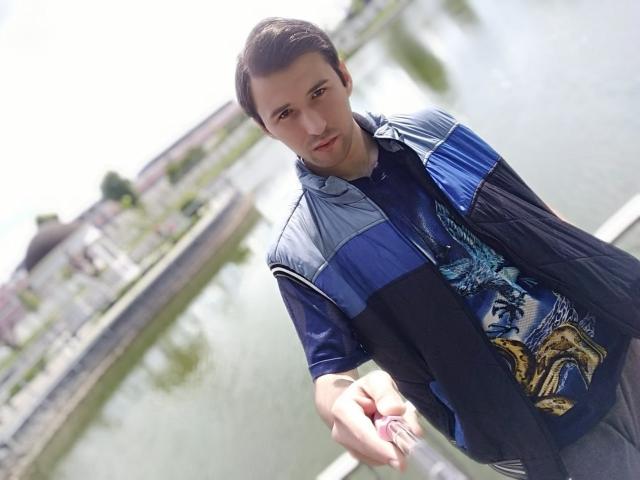 GregArdor, capelli marroni corti, giacca blu e nera, in piedi vicino all'acqua, che guarda in basso, mano destra tesa, sfondo leggermente sfocato, luce soffusa, aspetto casual, espressione leggermente seria..