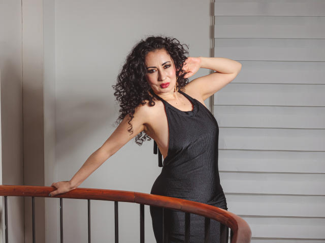 SamanthaAntolin with curly black hair, wearing a black tank top, leaning on a wooden railing, one arm raised, showing sideboob, standing in front of white blinds.