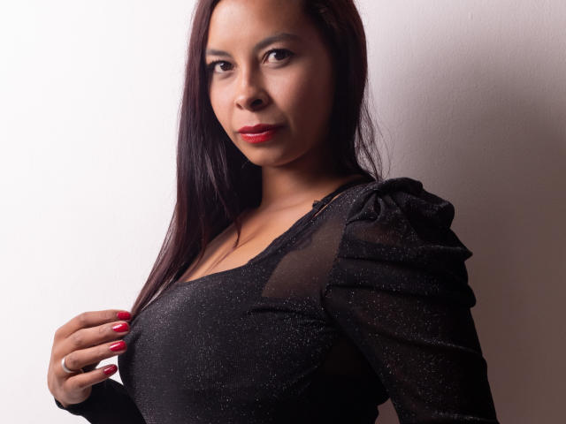 Clair Bennet, avec du rouge à lèvres, une robe noire pailletée, des manches bouffantes et des ongles rouges, touchant sa poitrine avec un sourire discret, devant un mur blanc uni..