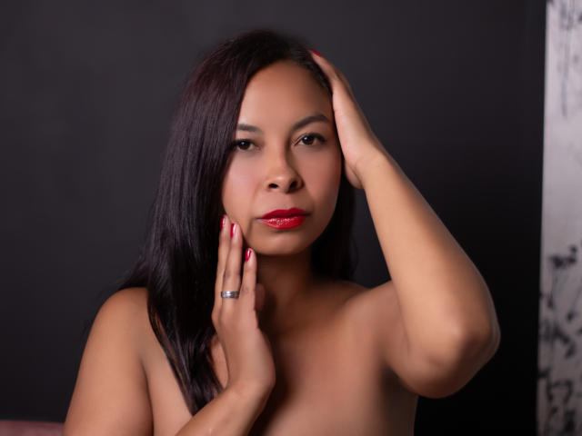 ClairBennet has dark brown hair, red lipstick, and a silver ring on her right hand. She's topless, with her left hand touching her head, showing off her tits.