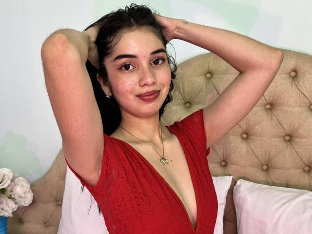 LunaCarter with arms up, wearing a red dress with a deep cleavage, showing her tits, smiling at the camera, long dark hair, light skin, and a necklace in a bedroom.