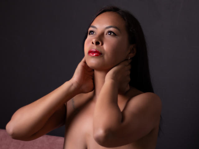 ClairBennet tiene la piel de color marrón oscuro, cabello negro largo, lápiz labial rojo y está sin ropa, con las manos tocando suavemente su cuello. Sus pechos son pequeños y su vulva no es visible..